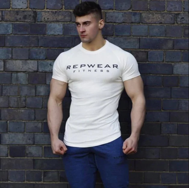 Athletic model in white REPWEAR FITNESS t-shirt poses against brick wall wearing blue pants, arms at sides