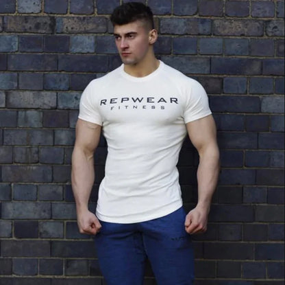 Athletic model in white REPWEAR FITNESS t-shirt poses against brick wall wearing blue pants, arms at sides