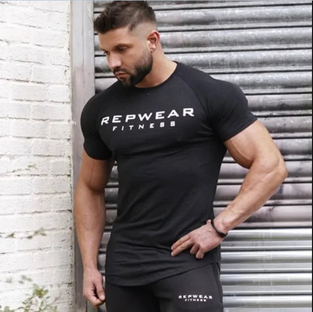 Athletic model wearing black REPWEAR FITNESS t-shirt poses against metal shutter, showing muscular physique