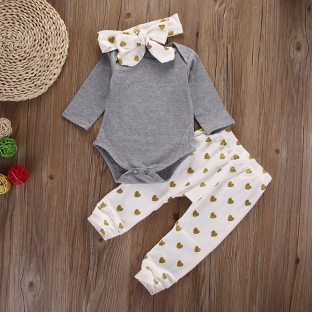 Baby outfit with gray bodysuit, white pants with gold heart pattern, and matching headband bow