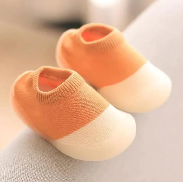 Coral orange knit baby socks with white rubber soles photographed on light gray textile surface