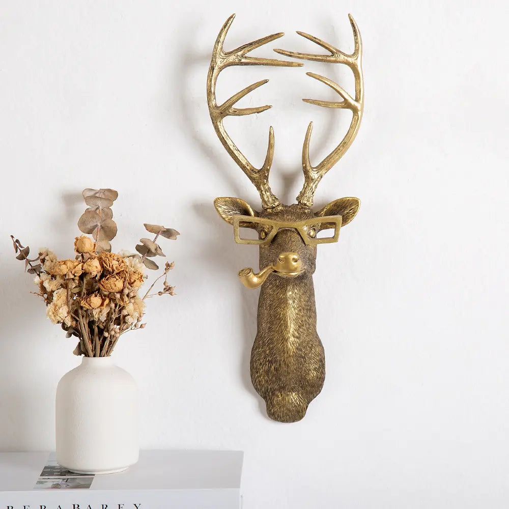 Golden deer head with glasses and a pipe, mounted on a wall next to a white vase with dried flowers