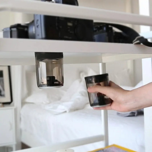 Hand placing or removing a transparent cylindrical speaker underneath a white shelf or desk, with a laptop visible in the background