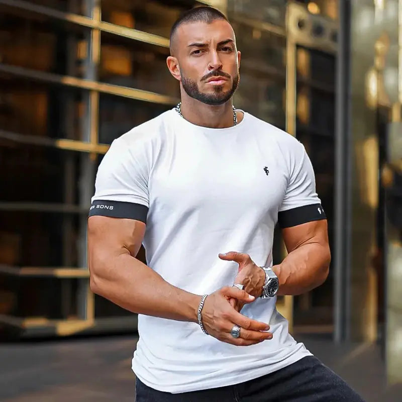 Man in white Father Sons t-shirt with black sleeve bands, wearing chain and watch, modern building backdrop