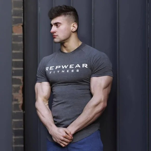 Model in gray REPWEAR FITNESS fitted t-shirt stands against dark wall, highlighting defined arm muscles