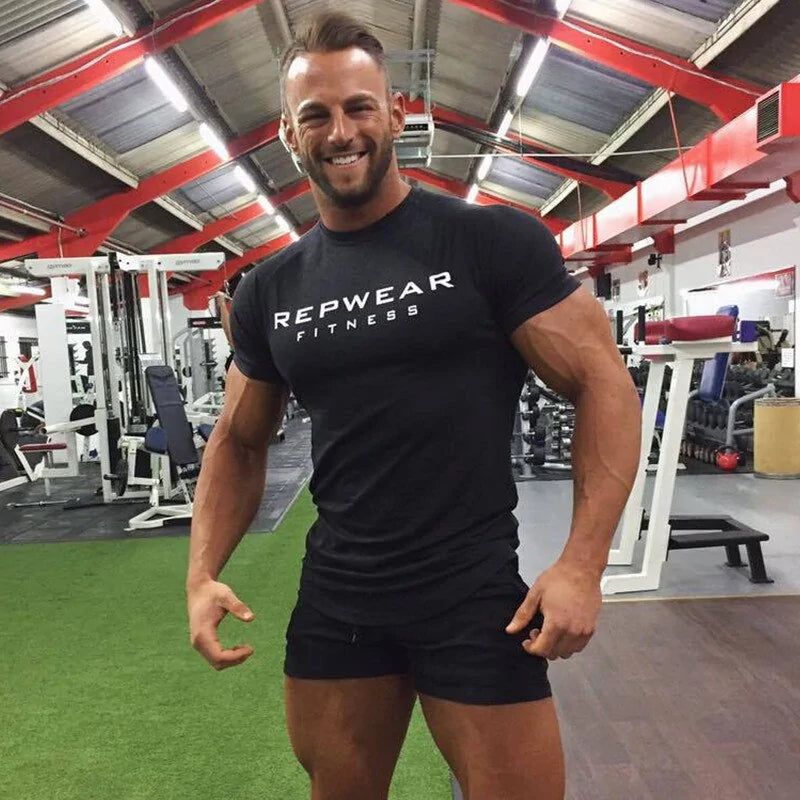 Muscular model in black REPWEAR FITNESS shirt smiling in gym setting with equipment and red ceiling beams
