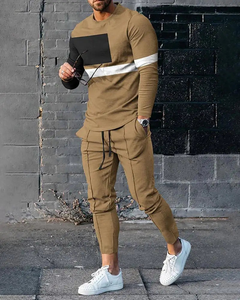 Person in tan tracksuit with black and white chest panels, jogger pants, and white studded sneakers against brick wall
