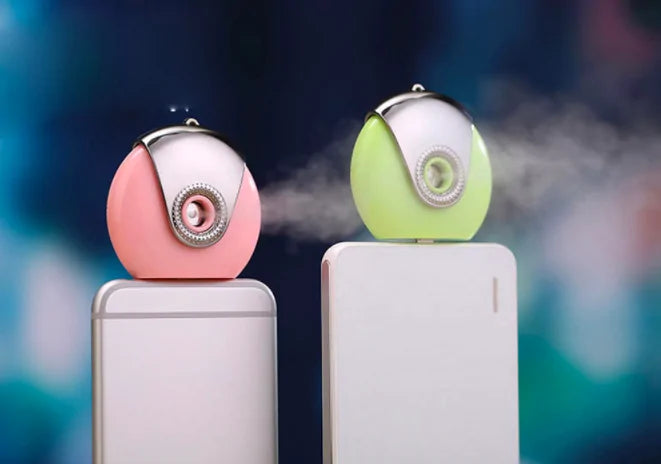 Pink and green portable facial misters emit vapor while attached to smartphones.