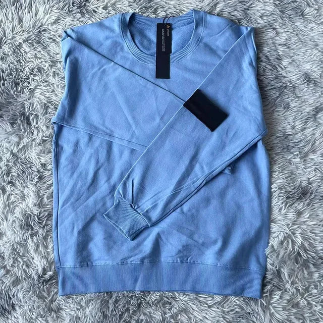 Sky blue cotton sweatshirt with black arm patch and price tag, displayed on gray textured surface
