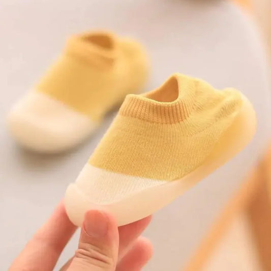 Soft yellow knit baby sock-shoe with white rubber sole, held by a hand against blurred background 