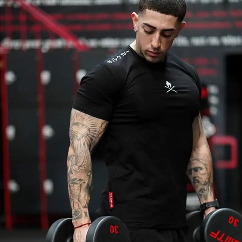 Tattooed athlete in black fitted shirt lifting dumbbells, logo visible on chest in gym setting