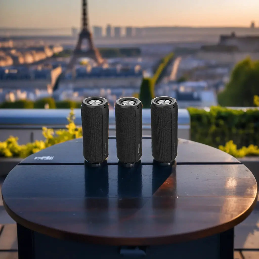 Three black cylindrical Bluetooth speakers on a round table with a cityscape and sunset in the background.