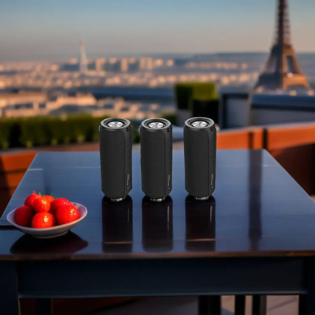 Three black cylindrical speakers on a table with a bowl of strawberries; blurred cityscape in the background featuring a tower.