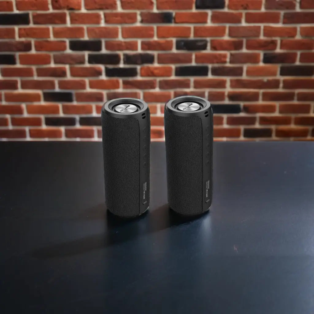 Two black cylindrical speakers with textured fabric, placed on a dark surface against a red brick wall.
