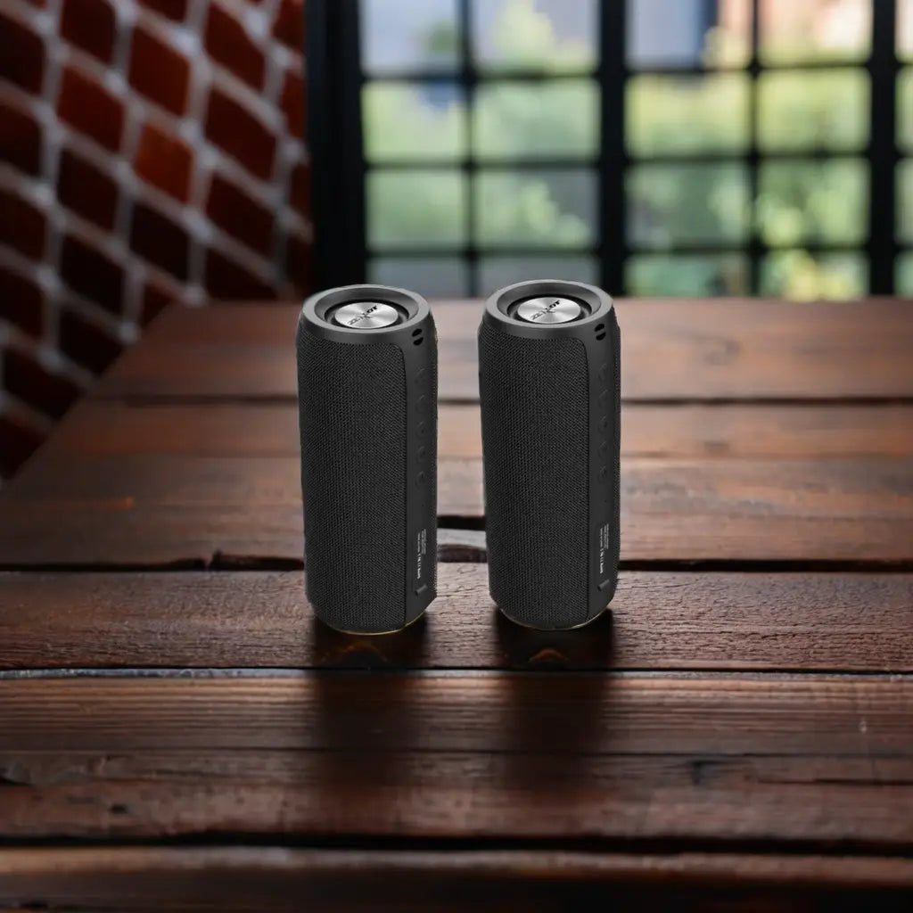 Two black cylindrical wireless speakers on a wooden table with a window in the background.
