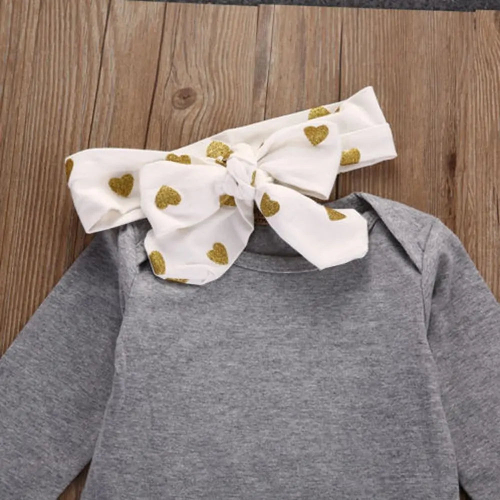 White headband with gold heart pattern and large bow attached to gray infant bodysuit collar