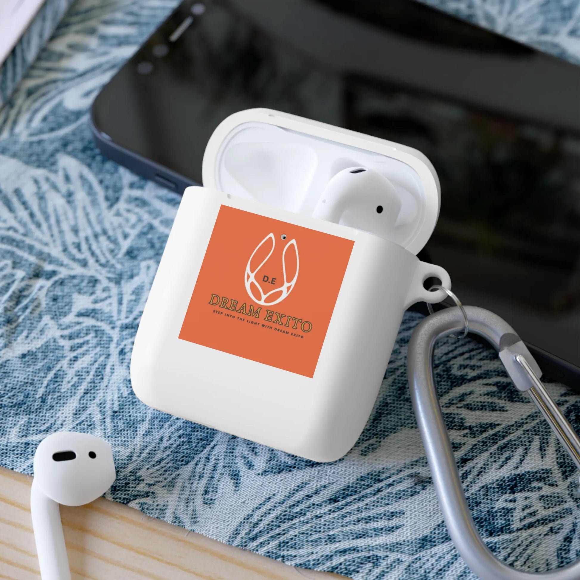 Wireless earbuds in a case with an orange "DREAM EXITO" logo sticker, on a patterned fabric